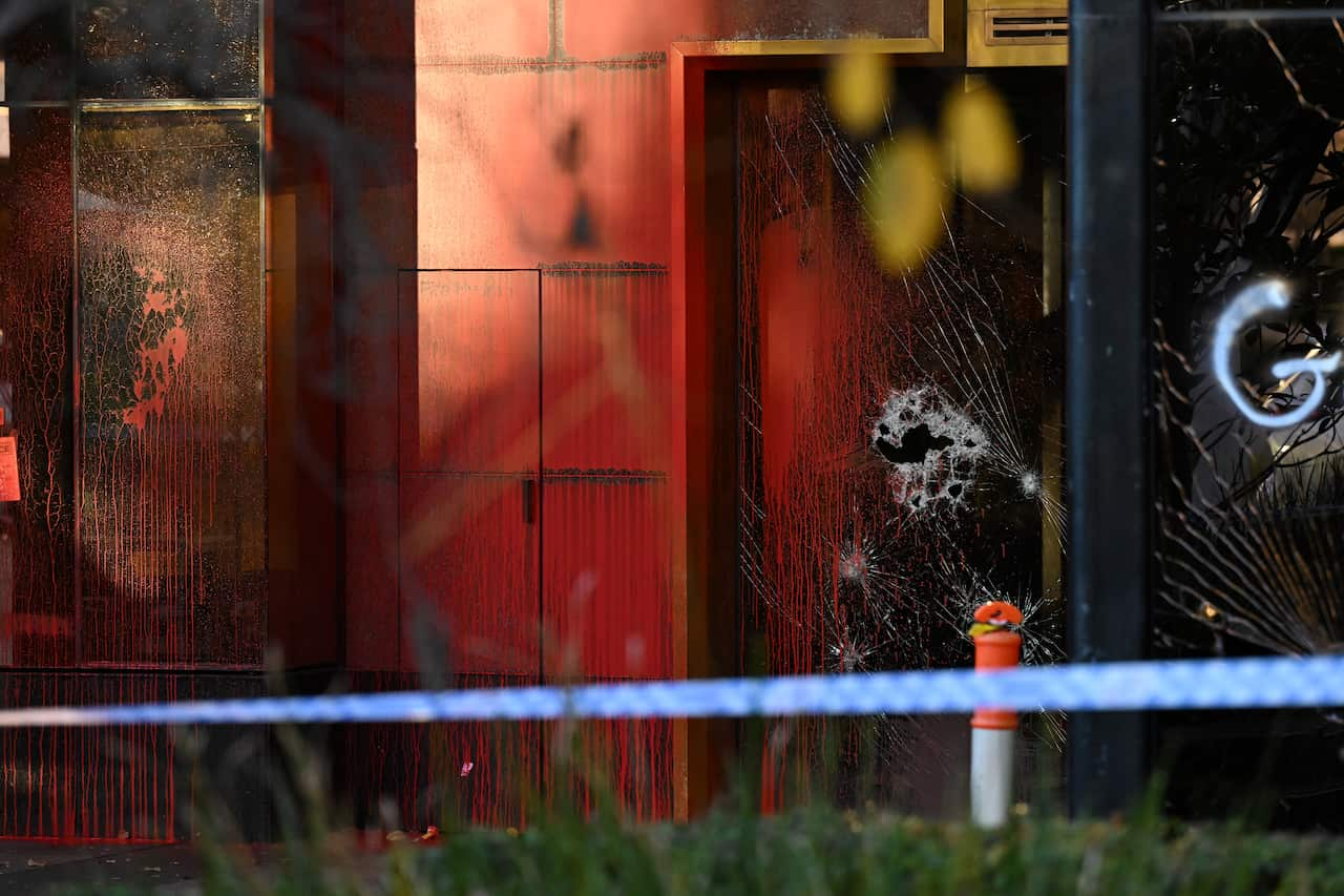 A building with smashed glass windows and red paint on it. Police tape is in front of it.