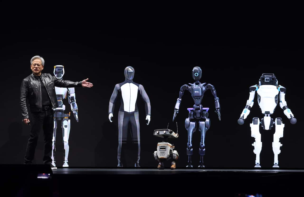 A man with grey hair and a leather jacket gestures towards several robots. 