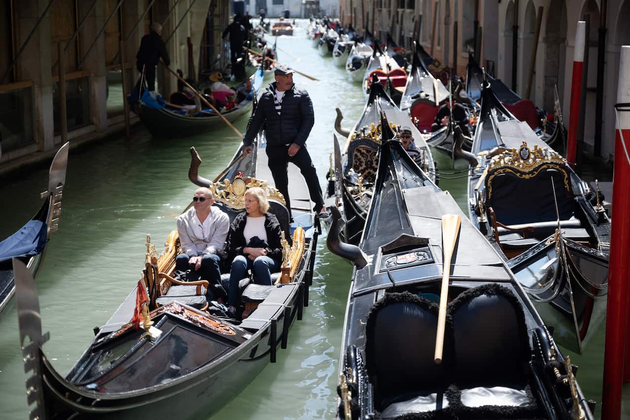 A gondolier in the venice, with two tourists onboard, navigates its way through a busy waterway,