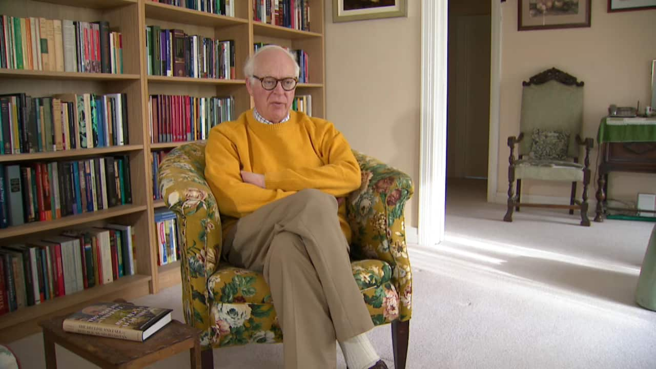 A man in a yellow jumper sitting in a chair with a flower print. There are rows of books on a shelf behind him.