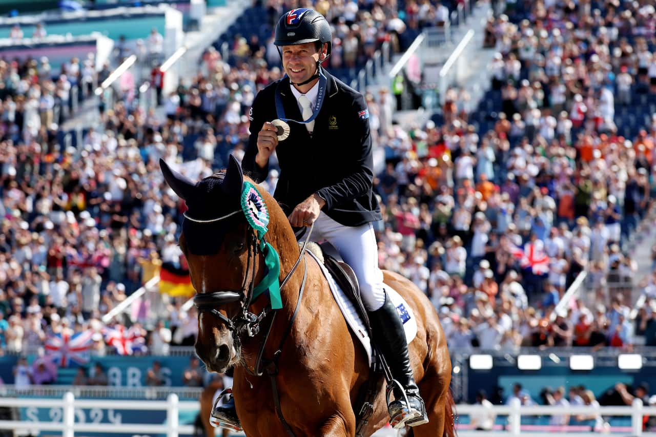 A man wearing a dark jacket, white pants, riding boots and helmet holds a medal in front of him while riding a horse