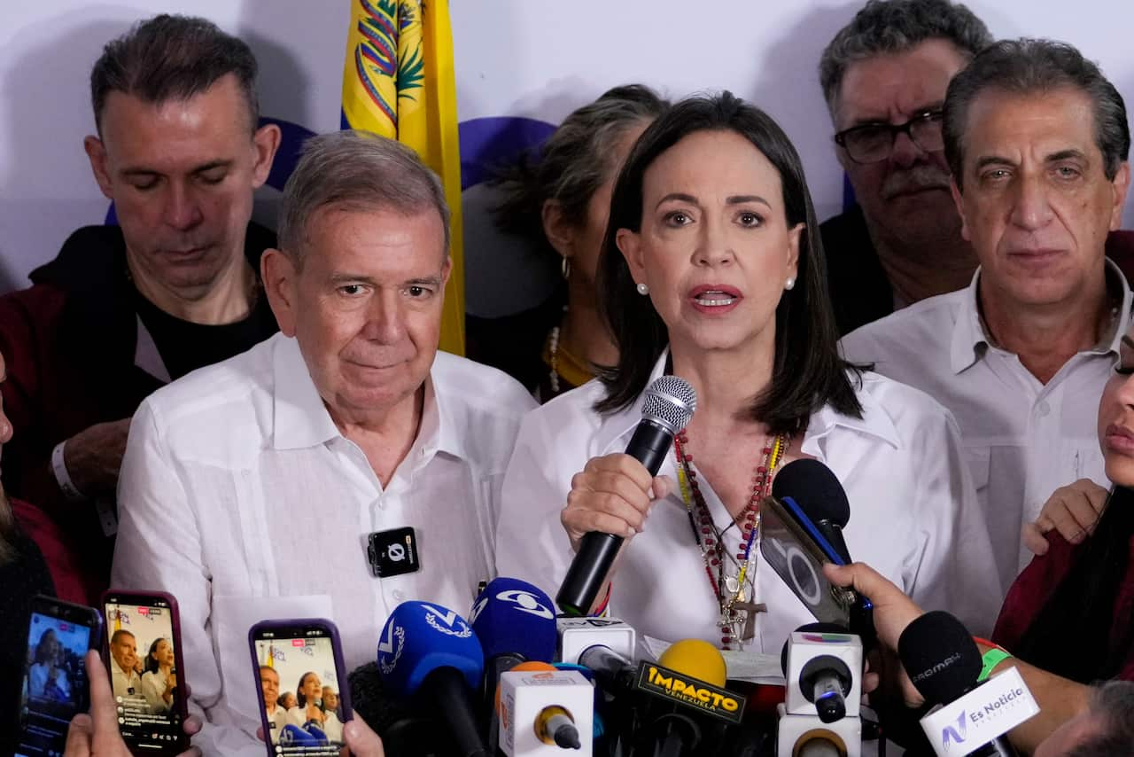 Edmundo Gonzalez Urrutia and Maria Corina Machado sitting at a table surrounded by media microphones.