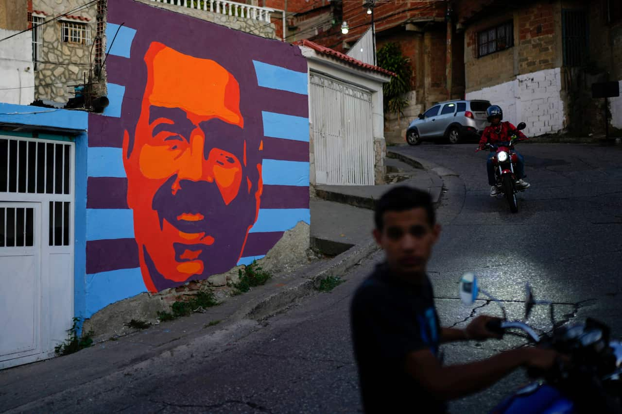 A street with a mural of Nicolas Maduro, with boys on motorcycles next to it. 