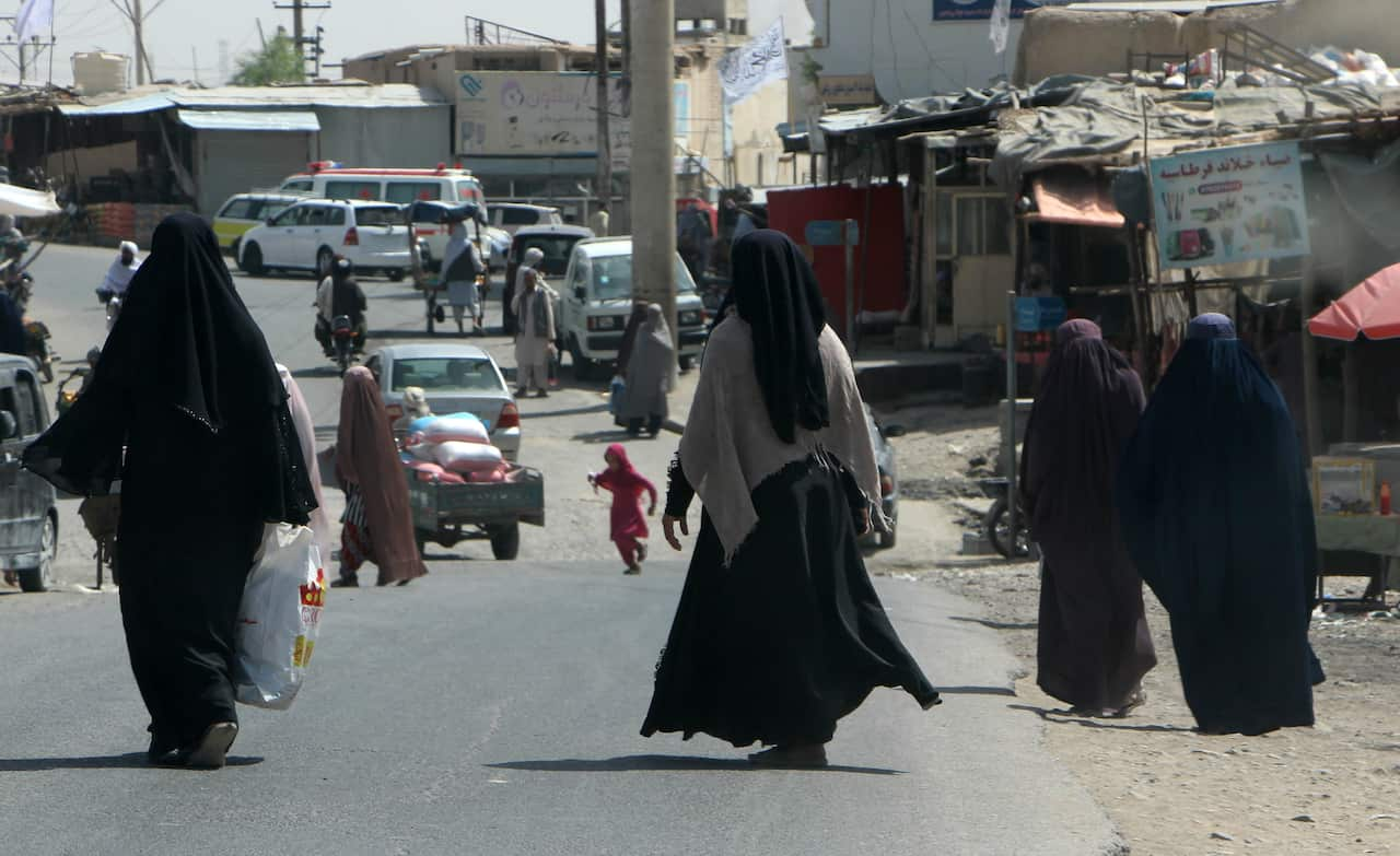 Burqa-clad Afghan women walking on a road. There are buildings and vehicles in the background behind them.