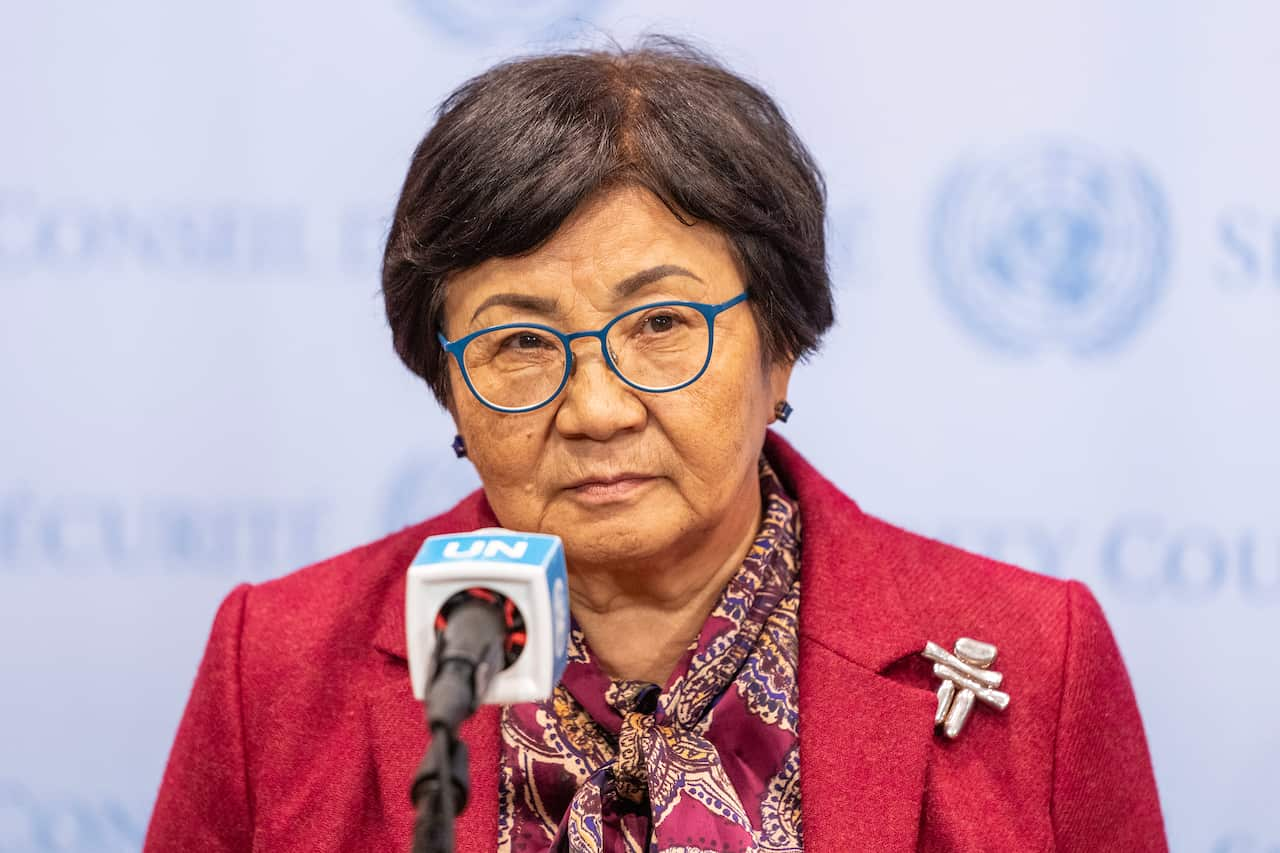 A woman wearing a red jacket stands behind a microphone that says "UN".