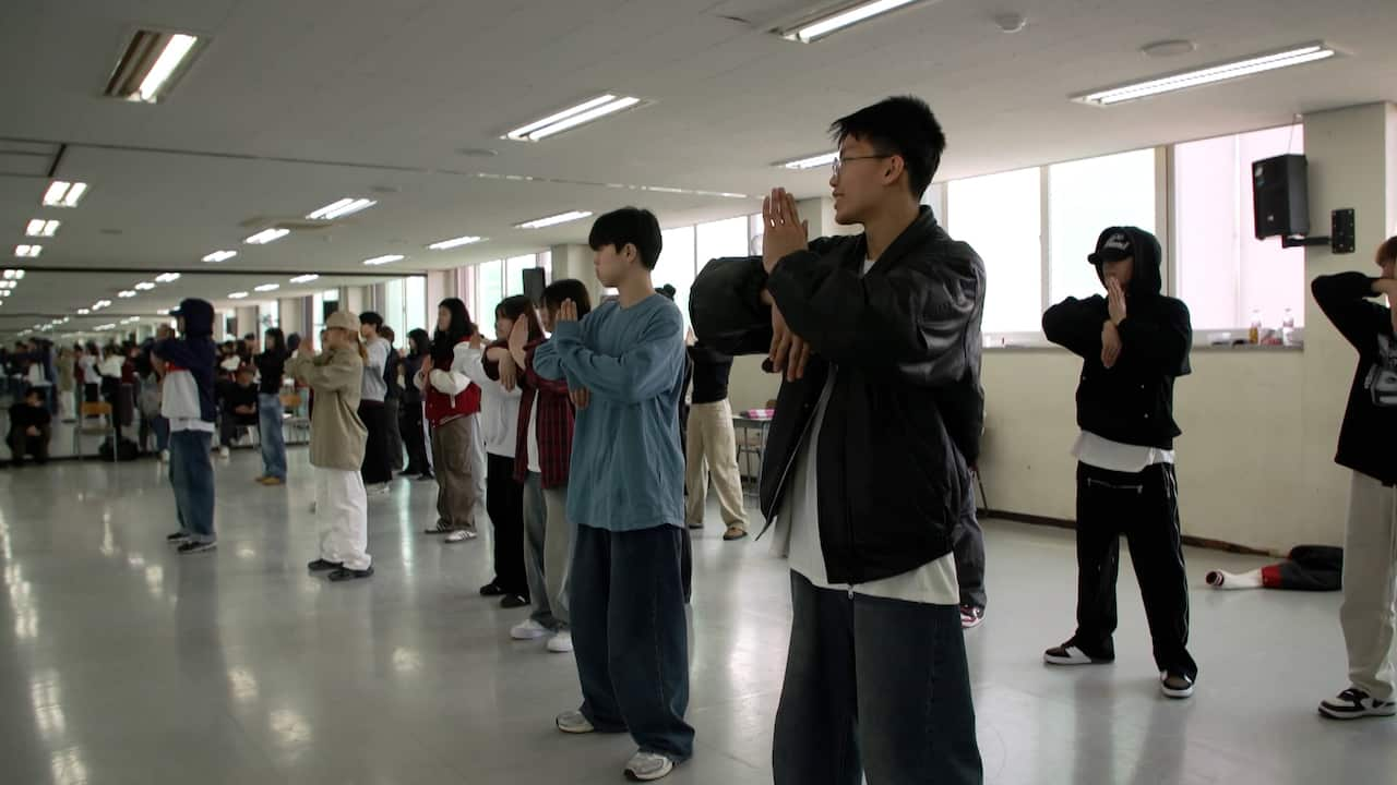 A group of korean K-pop dancers wearing baggy dark clothes stand in a line and hold their hands together, learning a new dance move. 