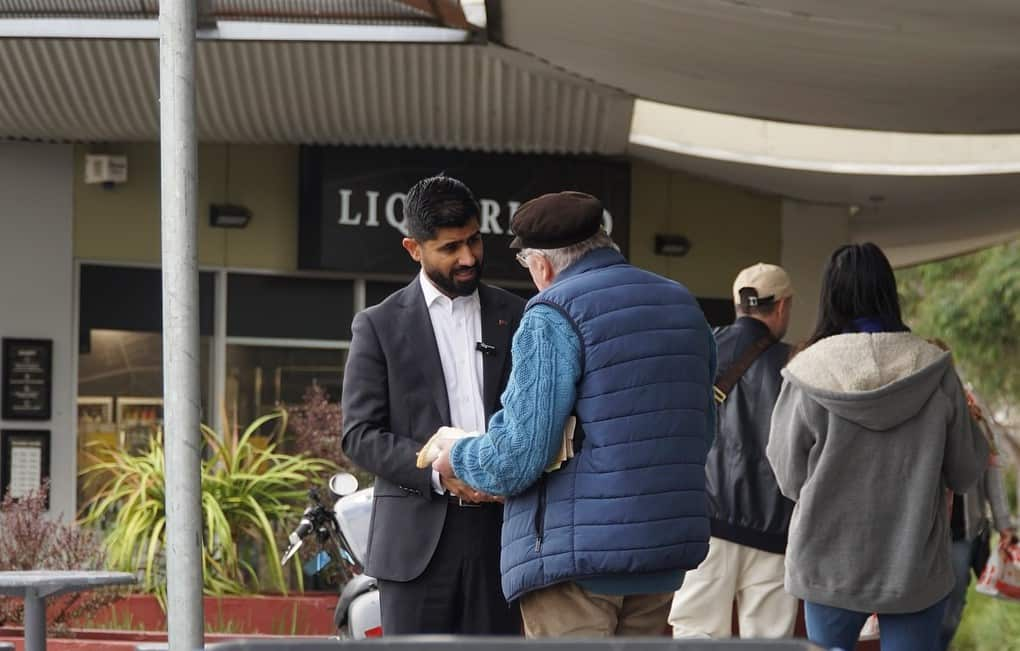 A man wearing a black suit and white shirt shakes the hand of an older man wearing a blue sweater near some shops. 