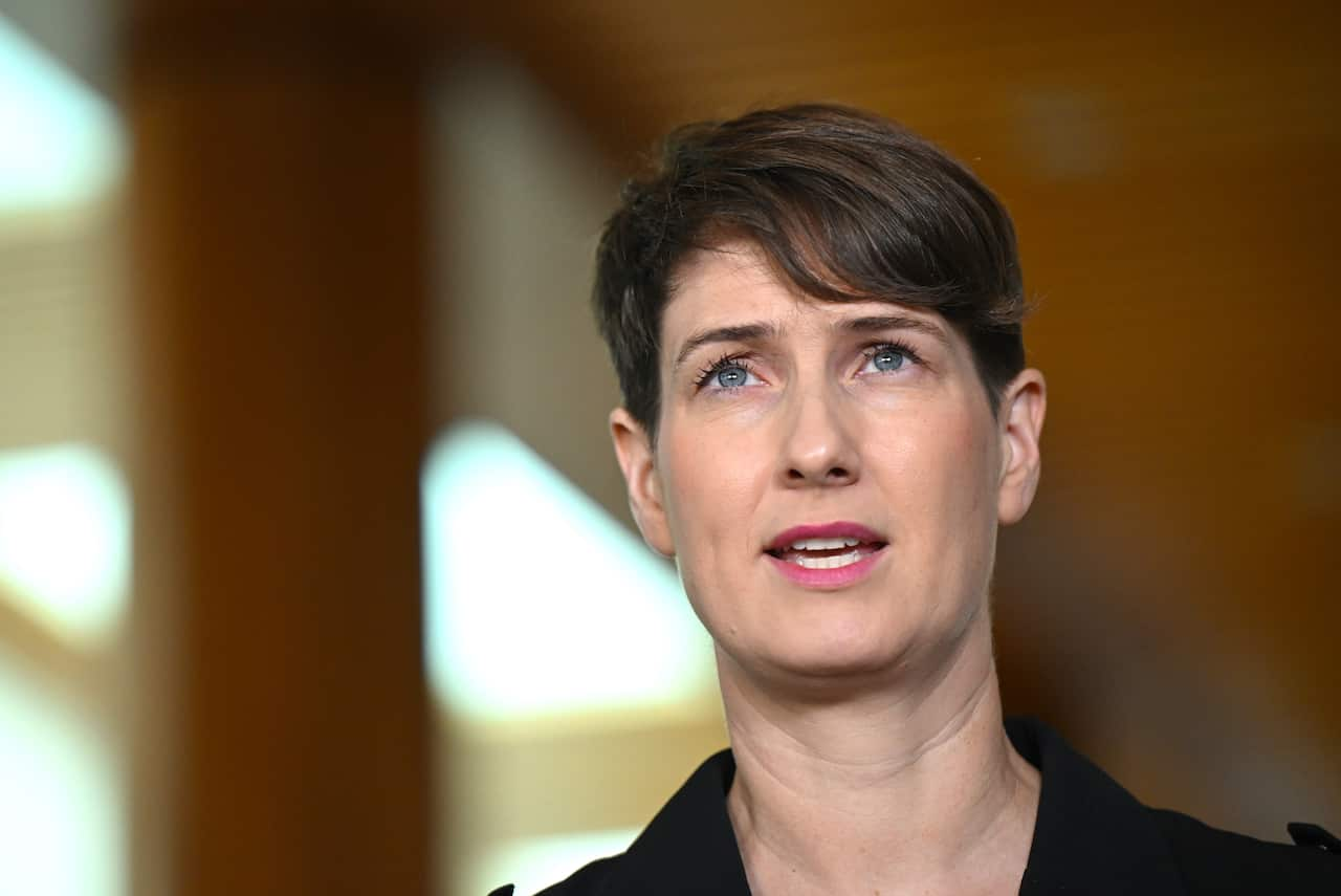 A woman with short hair wearing a black shirt is speaking at a press conference.