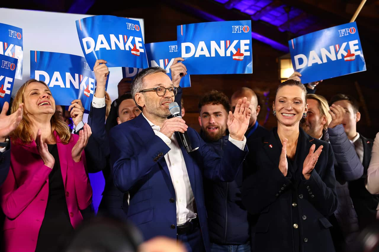 A man in a suit speaks into a microphone. He is surrounded by happy people. Some are holding up small blue signs reading danke.