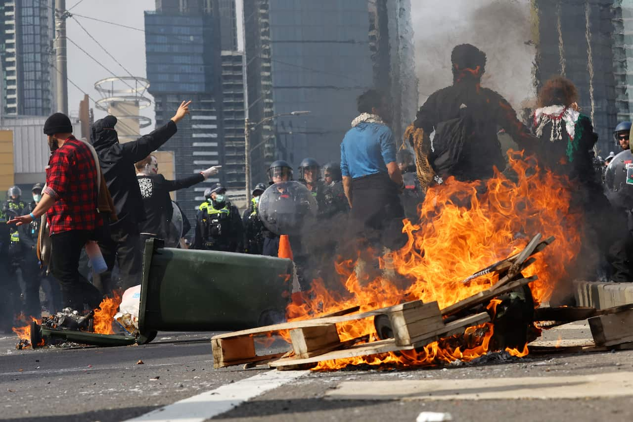 A pallet and bin shown on fire as protesters clash against police in the middle of a road.