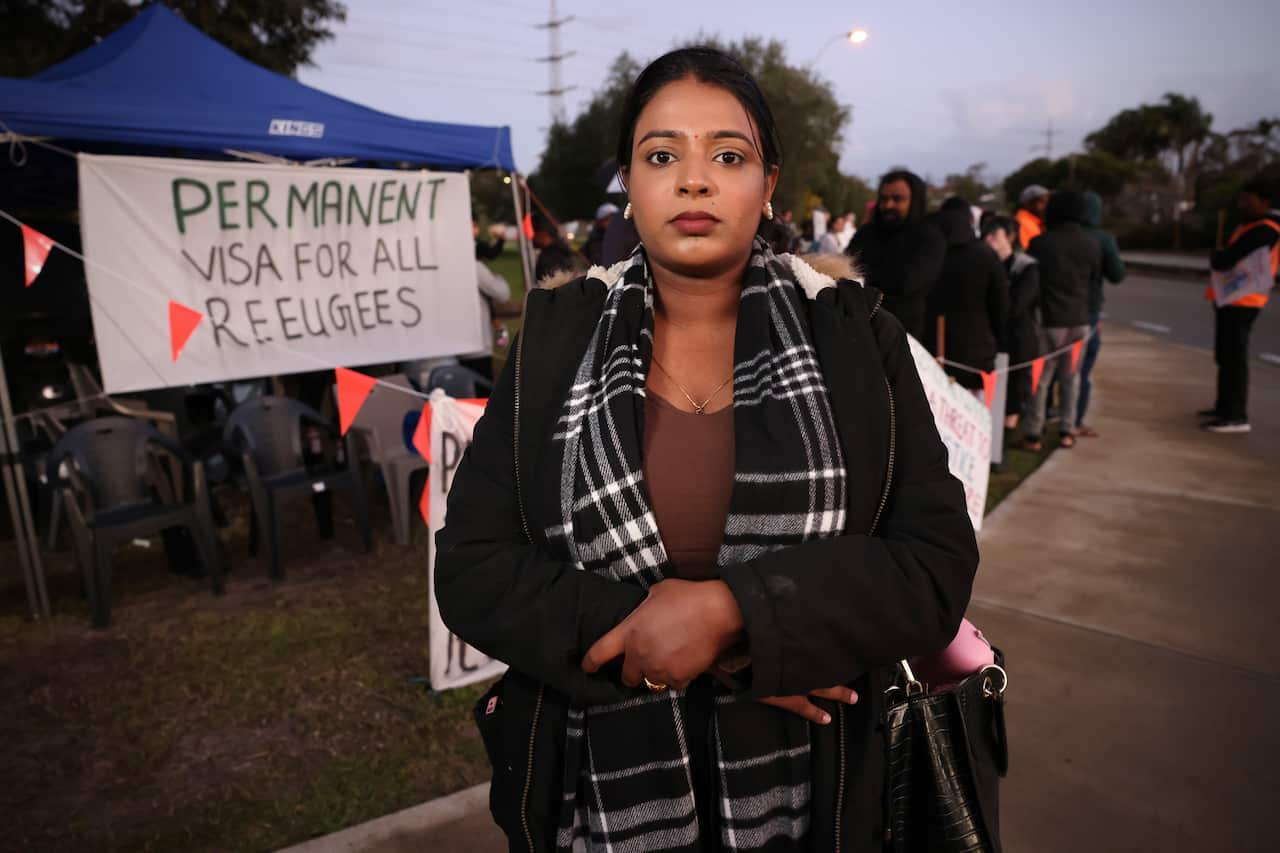 A woman standing in front of people and a sign that reads Permanent visas for all refugees