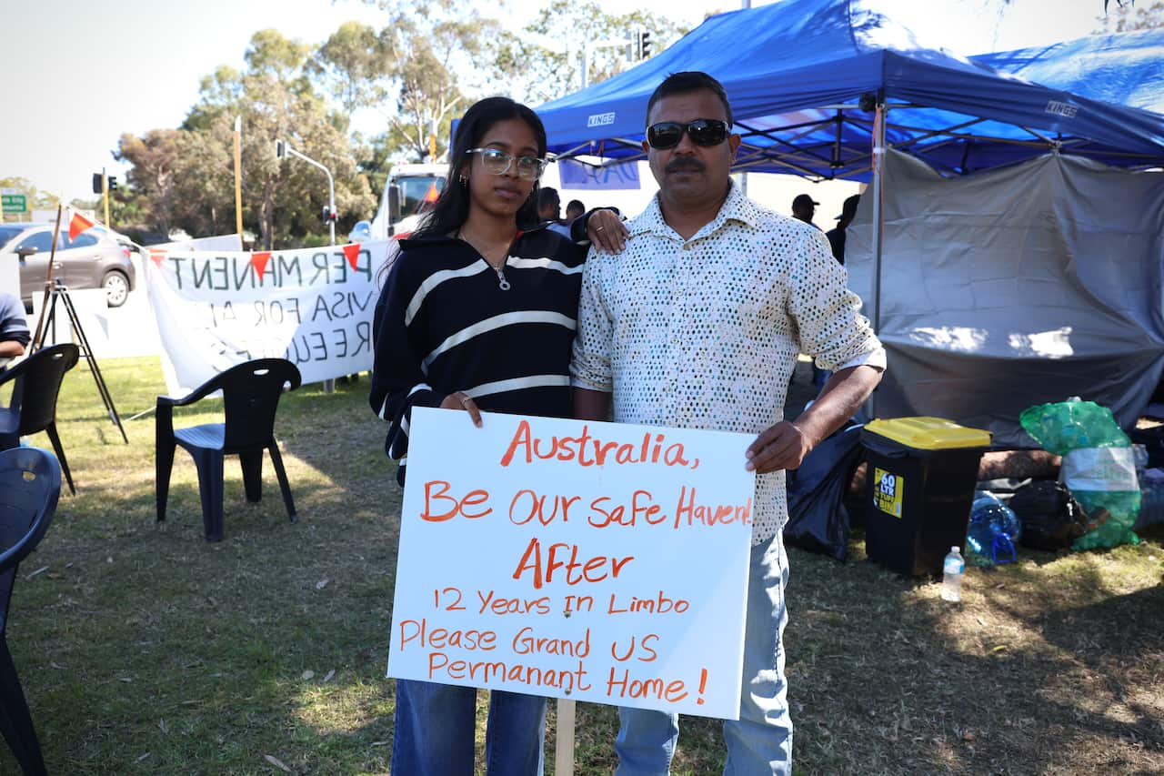 A man and a woman stand outside holding a sign in pleading for the federal government to grant them permanency in Australia after more than a decade on a visa.