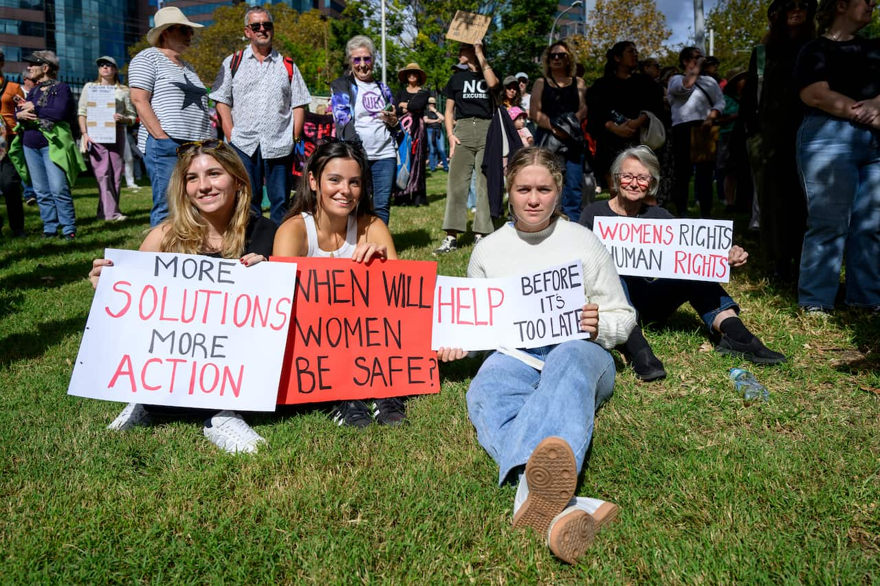 Four women, including three young women, sitting on grass with signs urging for stronger action against sexual violence.