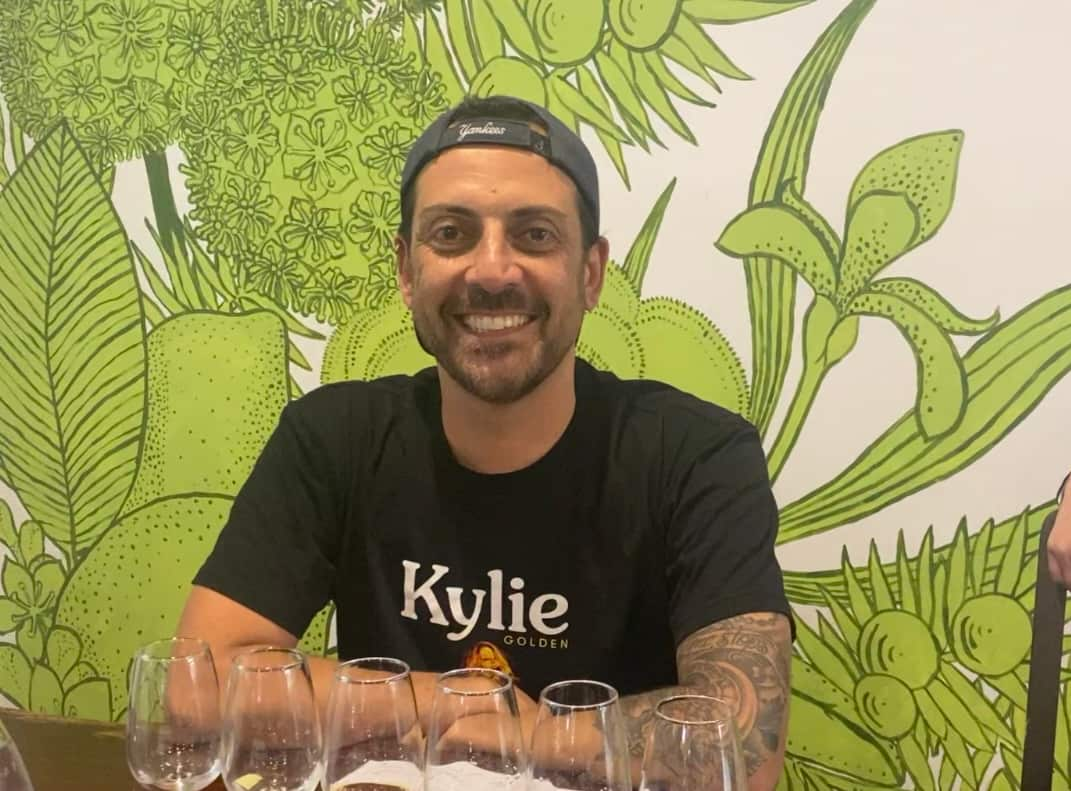 A man wearing a black t-shirt featuring the word Kylie in while letters smiles as he sits in front of a row of empty glasses.