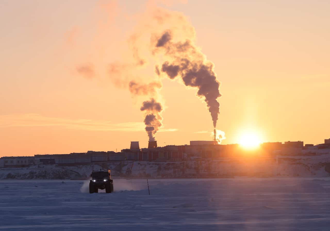 A jeep drives across what looks like snow with a low sun peeking out from behind an industrial facility releasing two large plumes of smoke. 