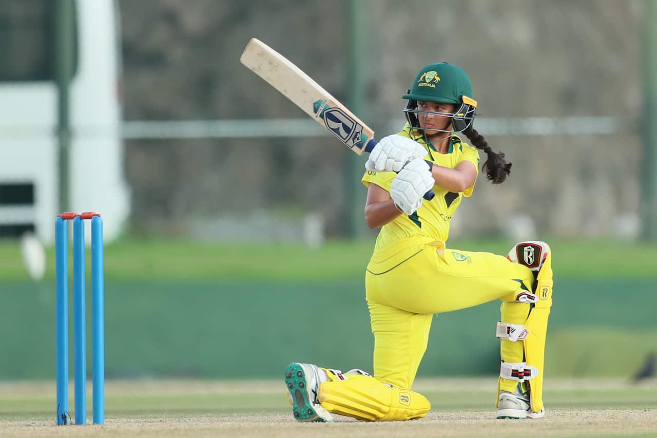 A woman wearing a yellow cricket uniform and a green helmet. She kneels and looks behind her after playing a cricket shot. 