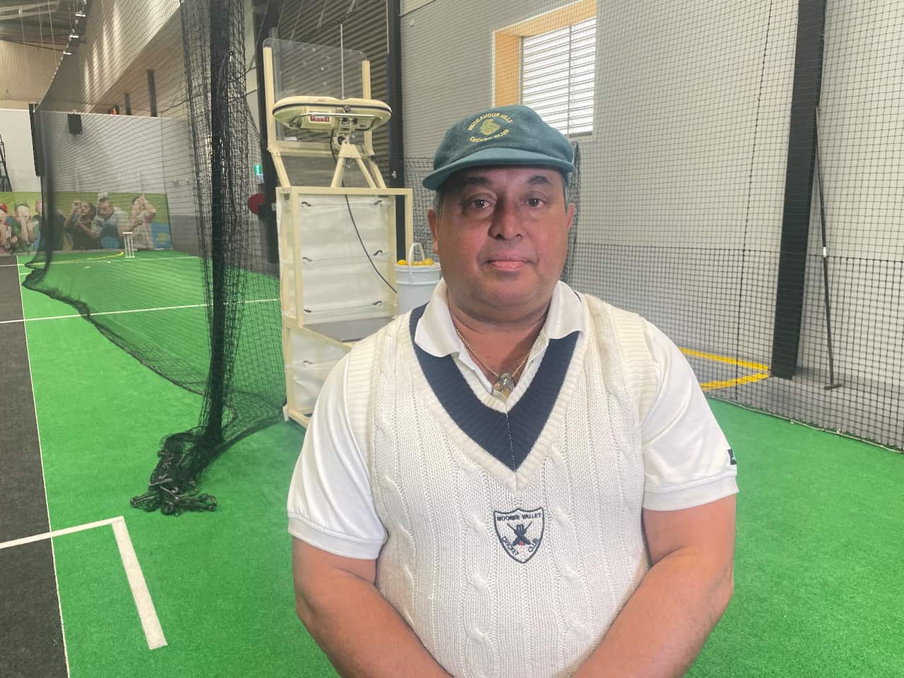 A man wearing a white cricket jumper and white shirt and a green cap posing for a picture in an indoor cricket net