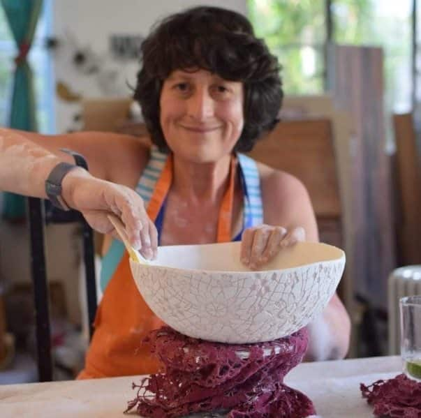 A woman holds a piece of incomplete pottery, her hands dirty with white clay, her face grinning out of focus in the background. 