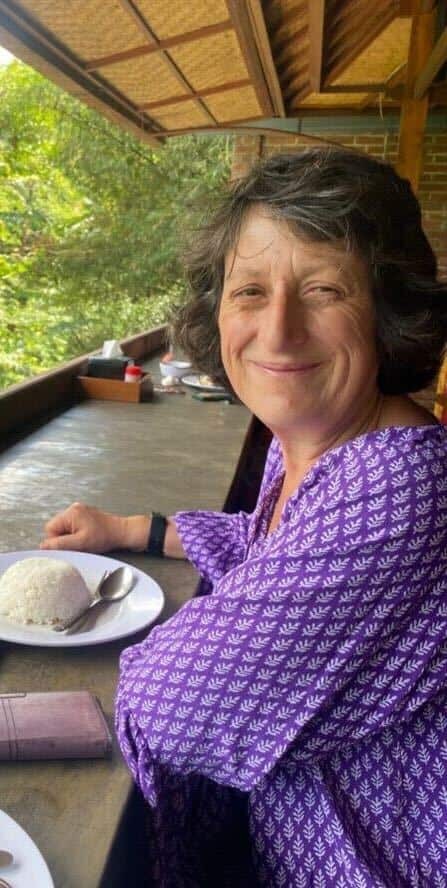 Greek Italian woman sits in a tropical location with a plate of white rice in front of her, grinning to the camera.