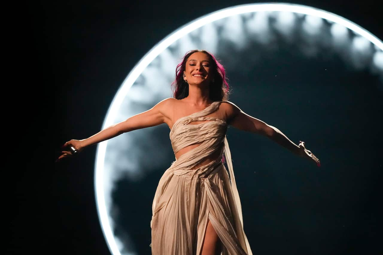 A woman wearing a white dress stands with her arms spread widely. A microphone is in her right hand. 