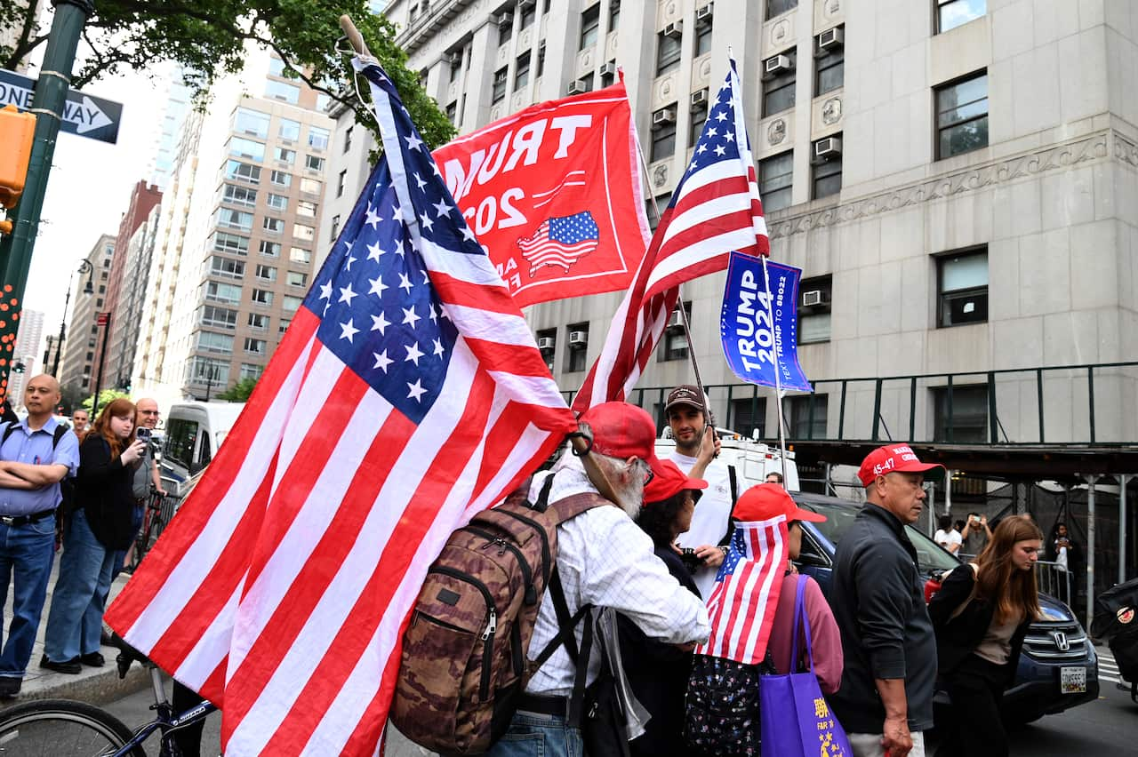 A group of people standing outside, holding American flags and some holding pro-Donald Trump banners.