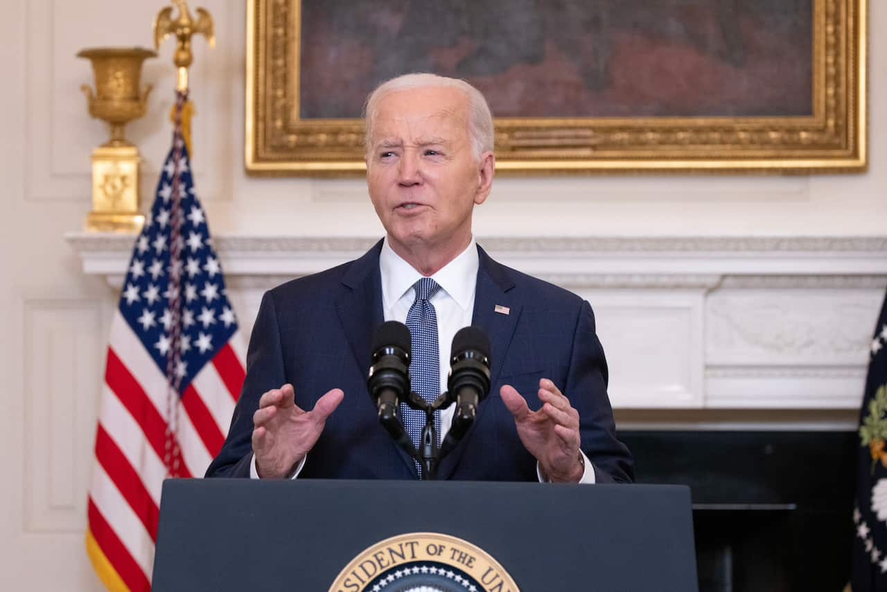 US President Joe Biden speaking into a microphone at a podium in front of a white wall and American flag.