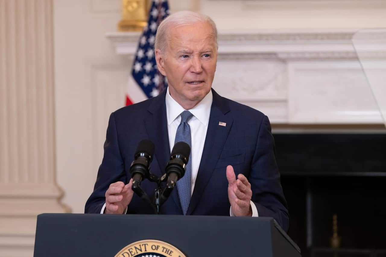 Joe Biden speaking into a microphone at a lectern. There is a white wall behind him with an American flag.