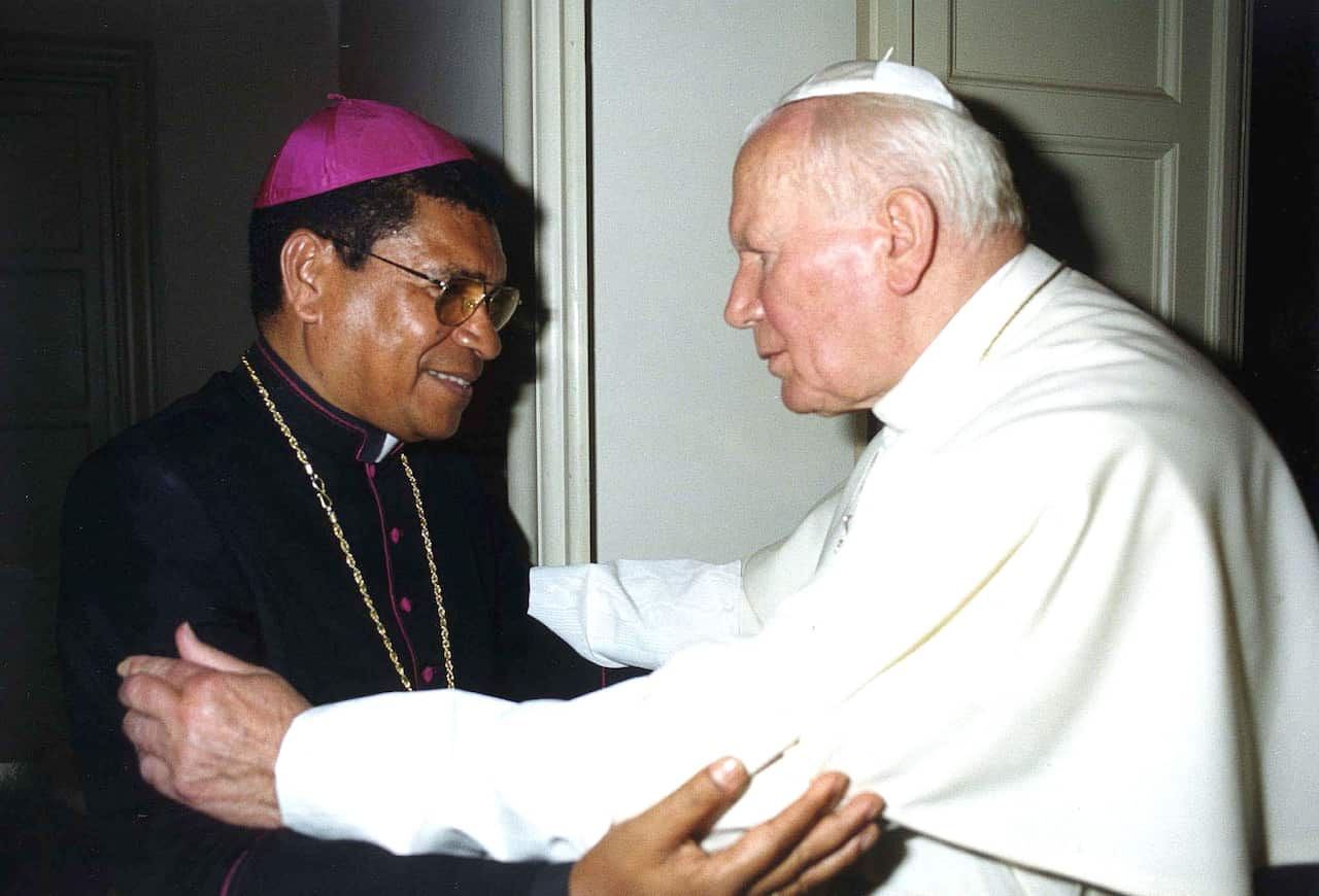 A man wearing a black clerical gown and purple cap (left) embraces Pope John Paul II, who is wearing white.