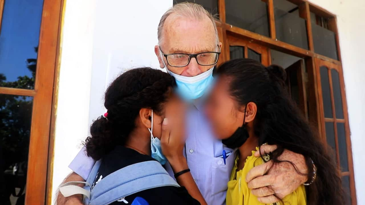 A bespectacled older man wearing a medical face mask hugs two young women whose faces are blurred 