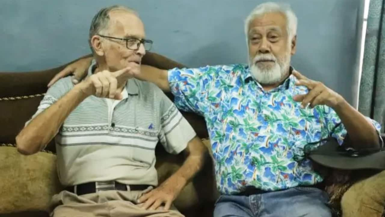 Two older men sitting on a couch. The man on the right is wearing a floral blue shirt and has his right arm around the shoulder of the man on the left, who is pointing his index finger 