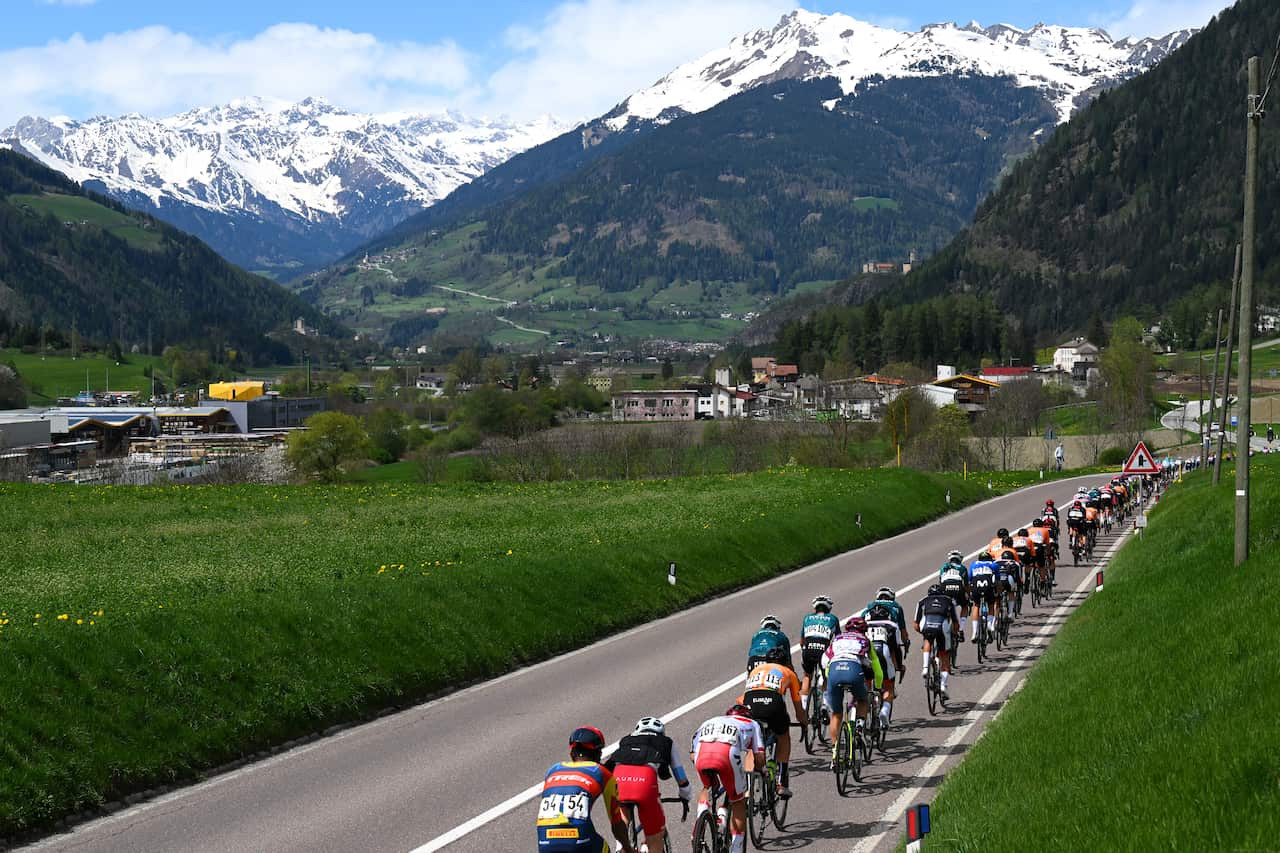 Riders pass through a small town with snow-capped mountains in the background.