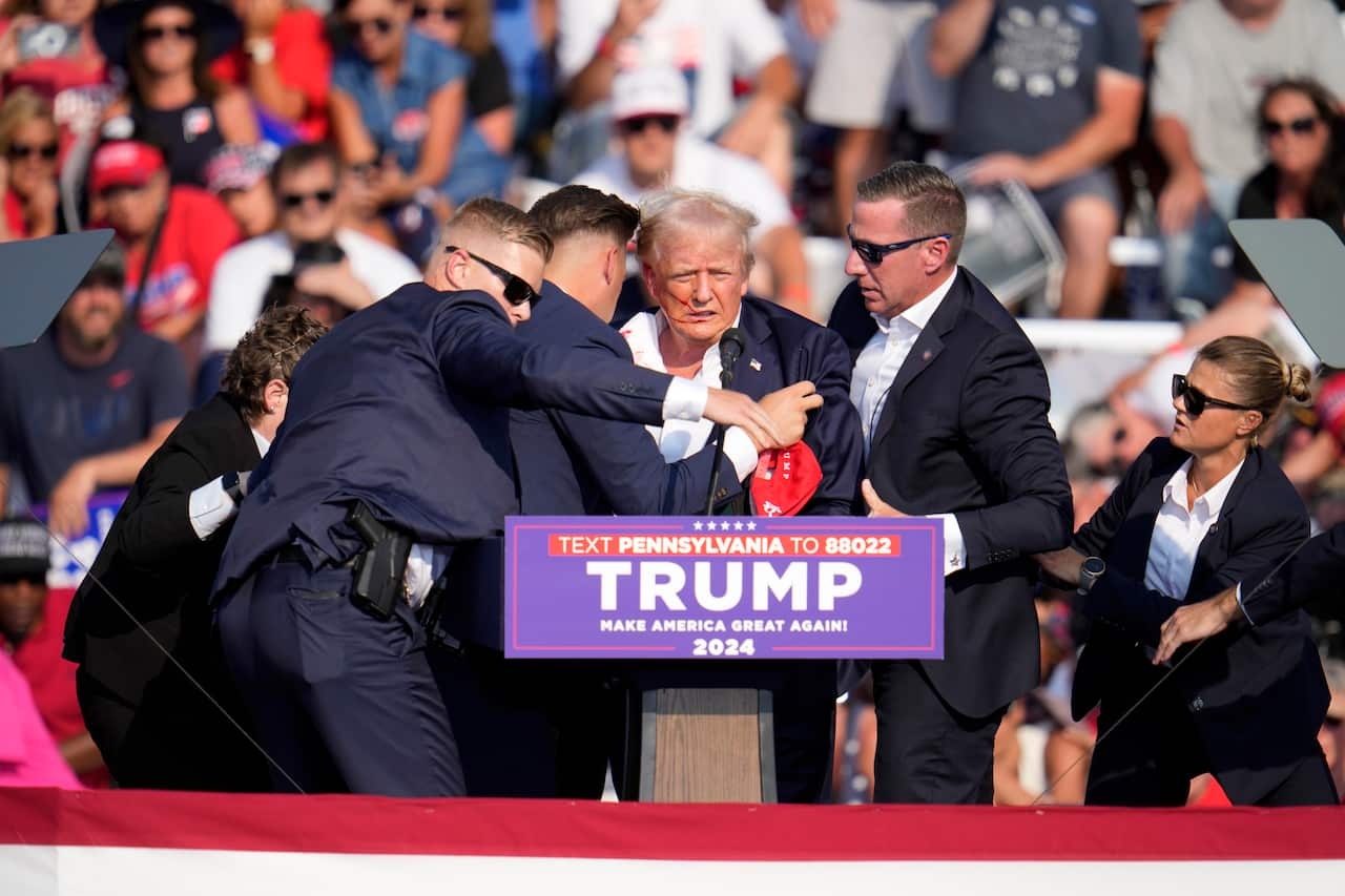 Donald Trump being shielded by men in suits. Audience members are behind him on bleachers.