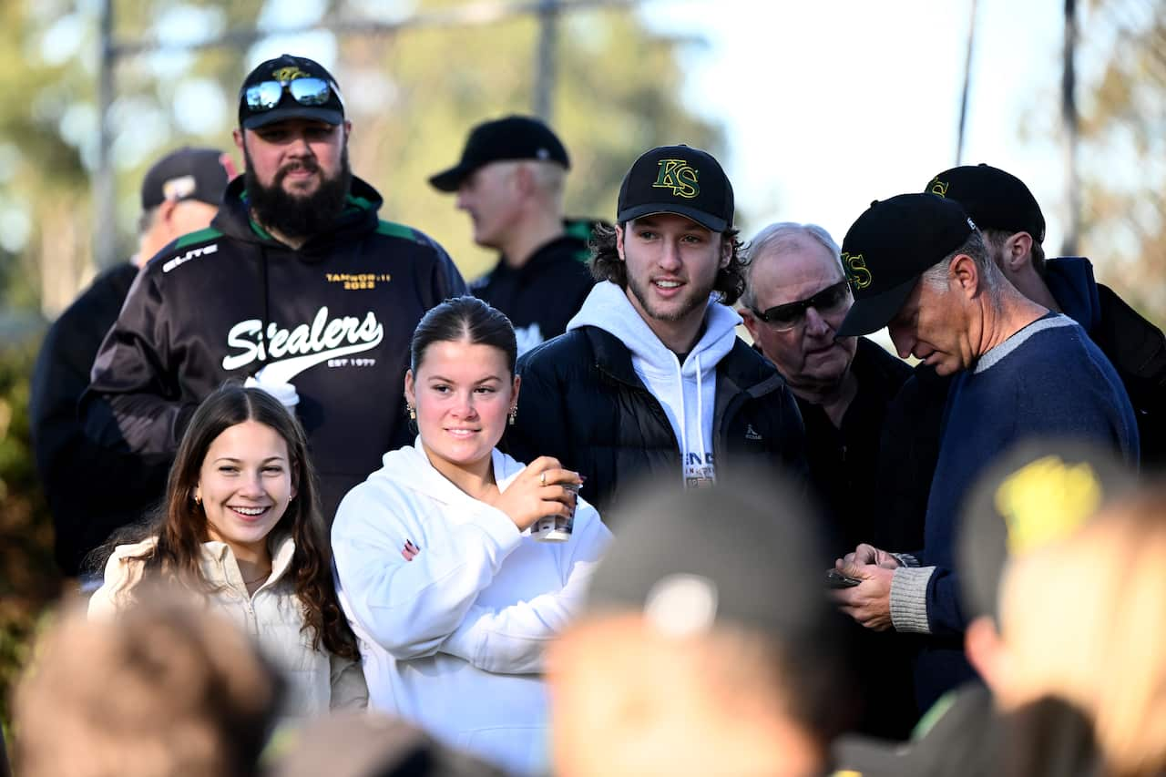 A crowd of people stand around. Some are huddled looking at a phone.