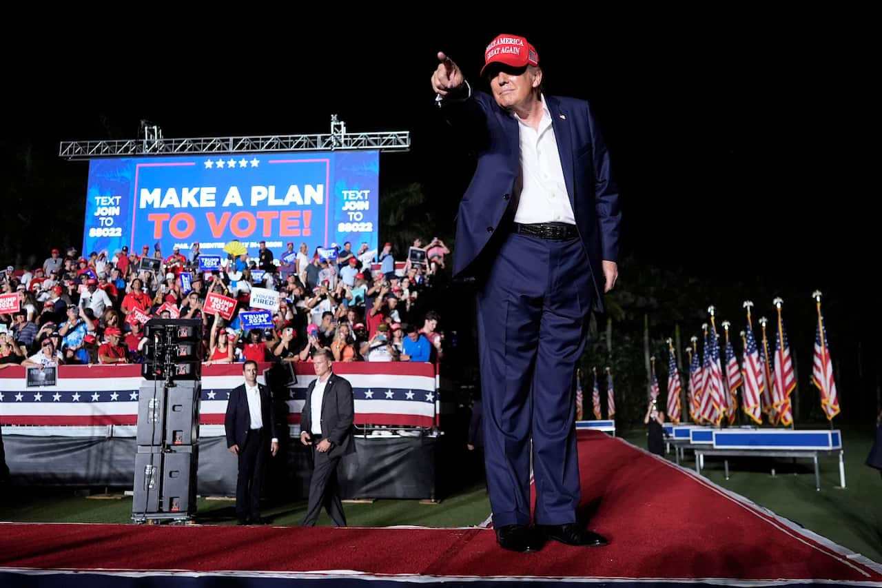 A man wearing a red cap and blue suit points at audience from a stage.