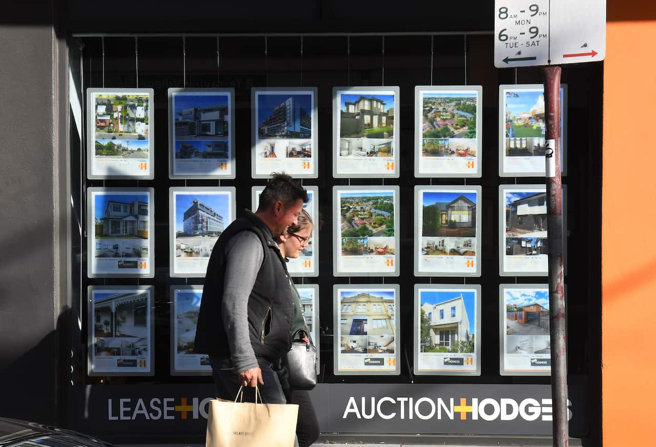 A couple walk past a real estate agent's window with home listings displayed on it.