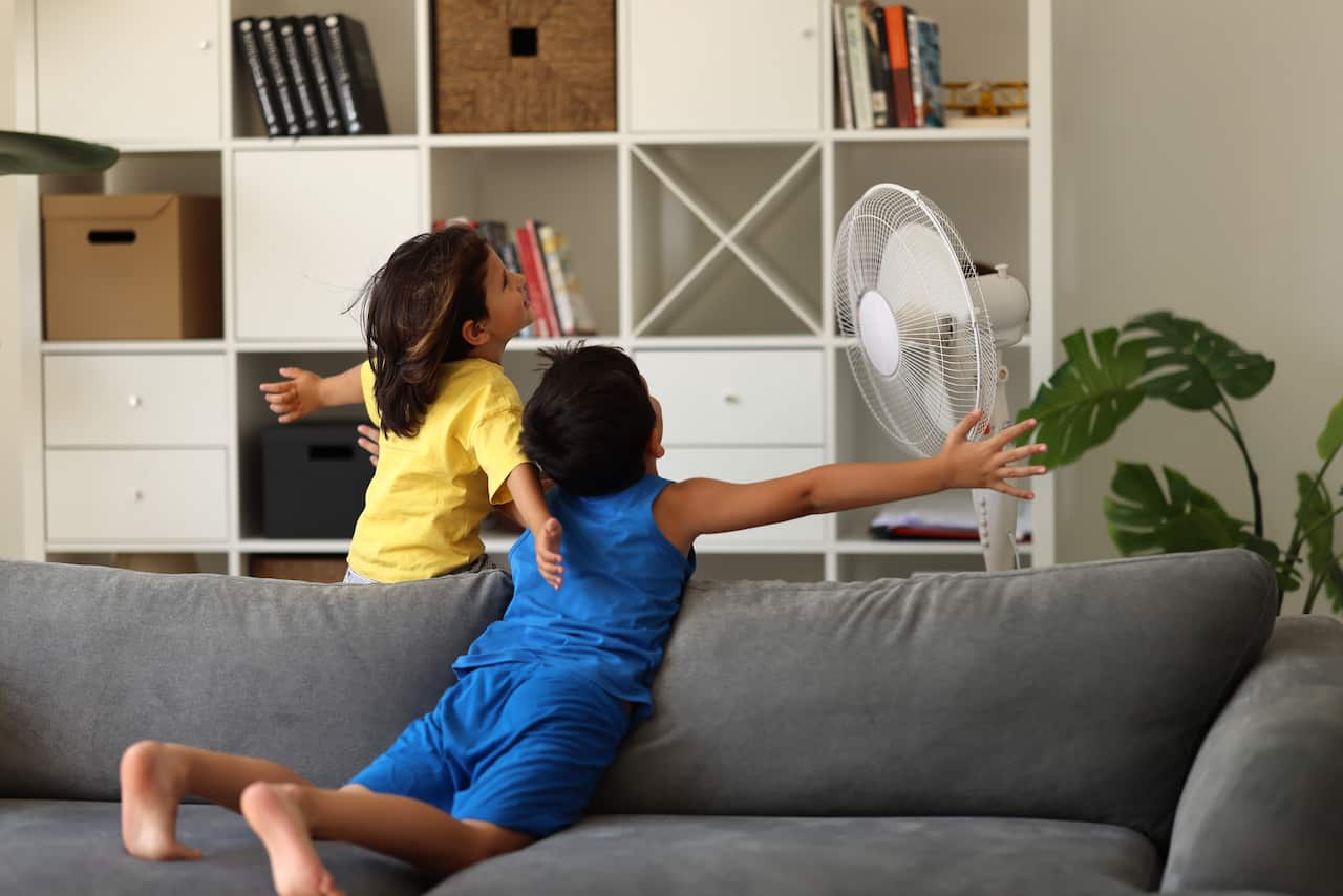 Children standing by a fan inside a house.