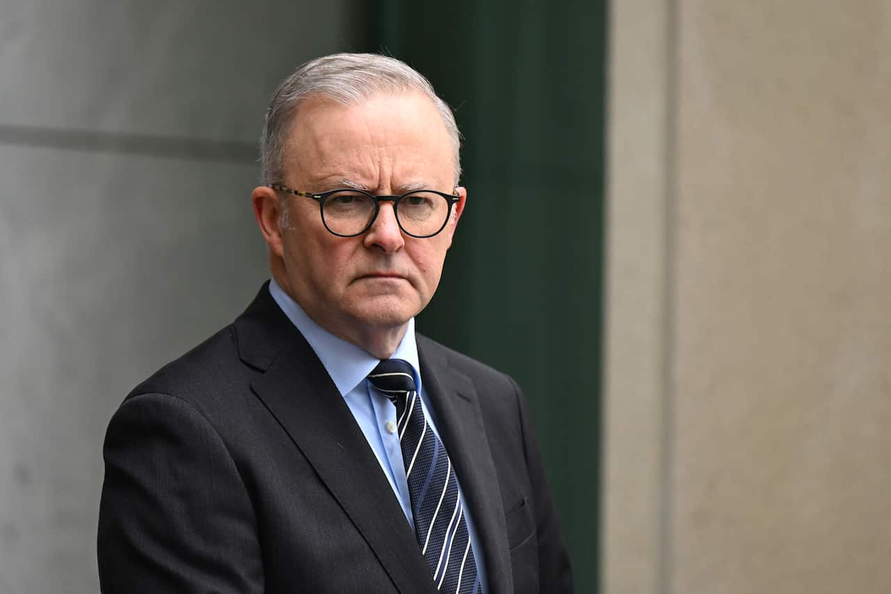 Anthony Albanese wearing a suit, tie, and glasses listening while at a press conference that is behind held outdoors.