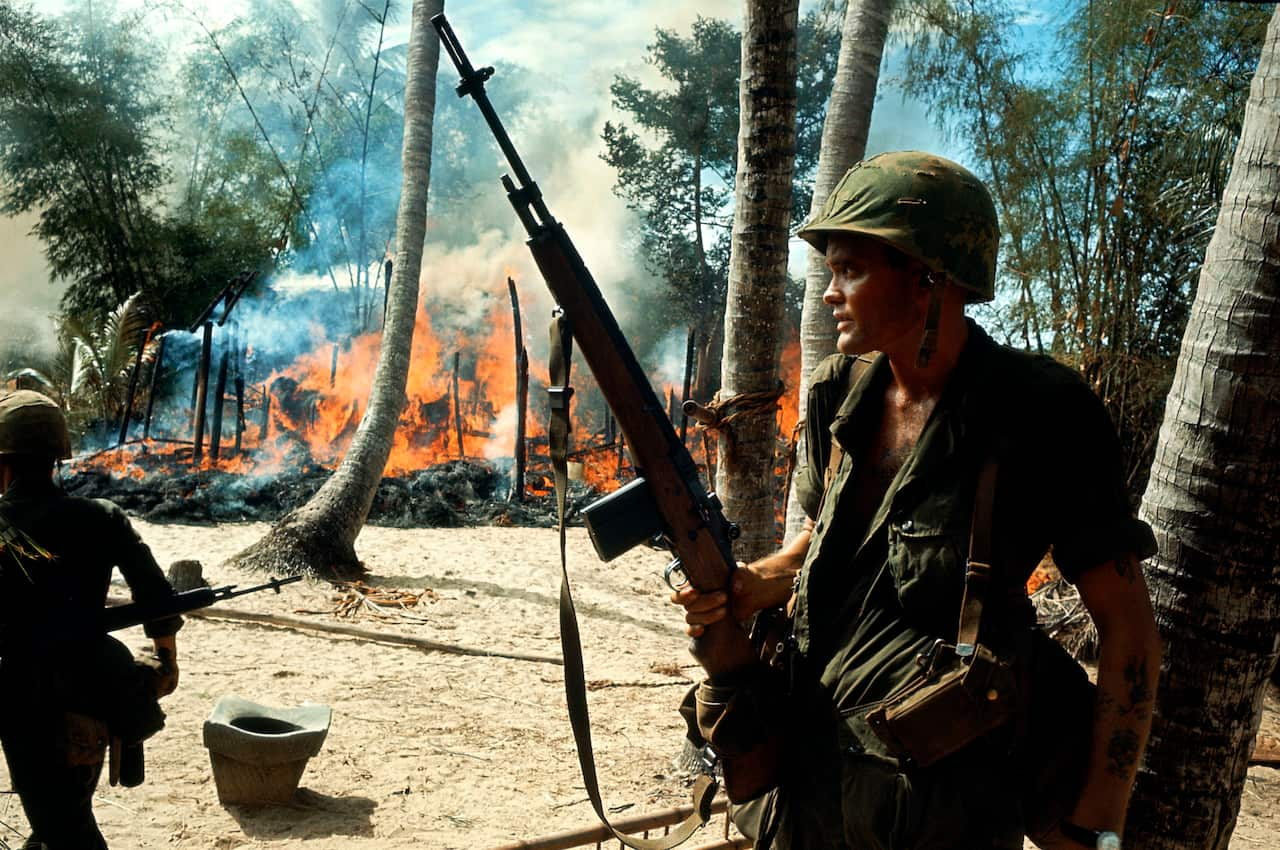 A soldier in green stands before tree trunks and a burning building in the near distance 