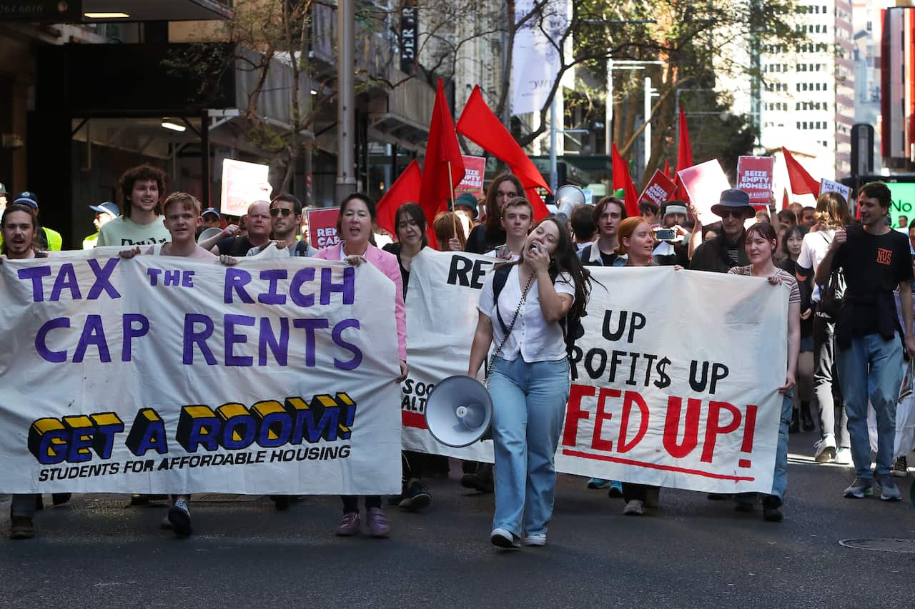 People marching in the street. A woman is holding a loudspeaker and some people are holding a banner reading: Tax the rich cap rents. 