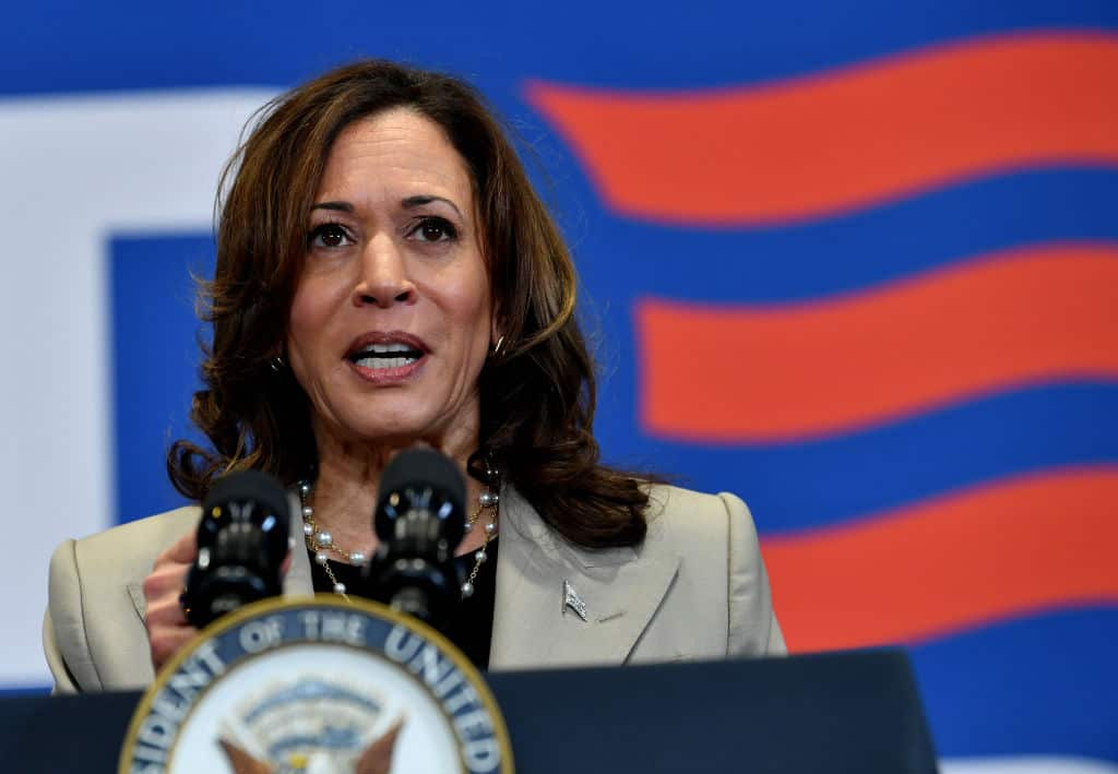 A woman stands at a podium with a microphone, with the US flag in the background.