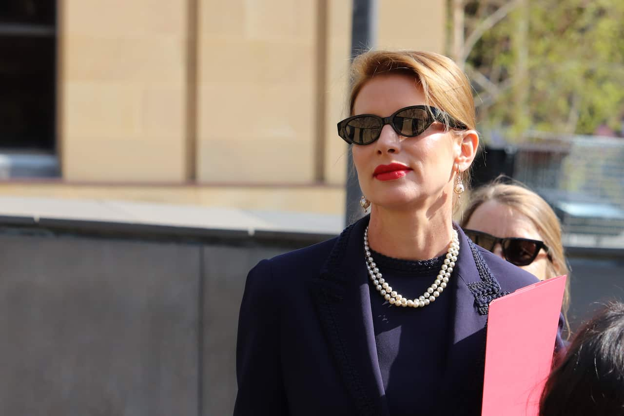 A woman wearing black sunglasses, a pearl necklace and holding a red envelope outside a building.