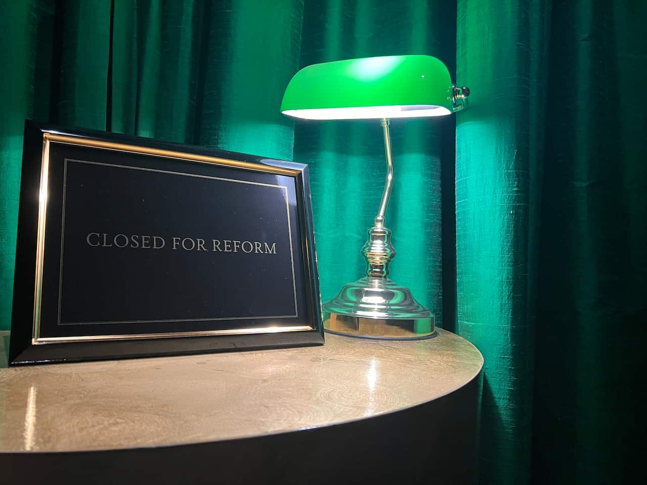 A table with a picture frame that says "Closed for Reform", next to a lamp. A green curtain is behind the table and objects.