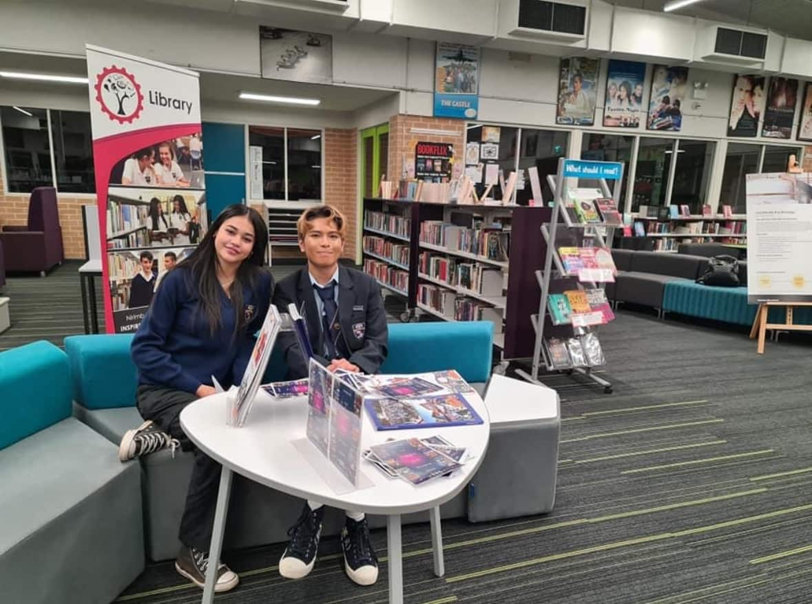 Two school students posing for a photo while seated on a bench in a library.