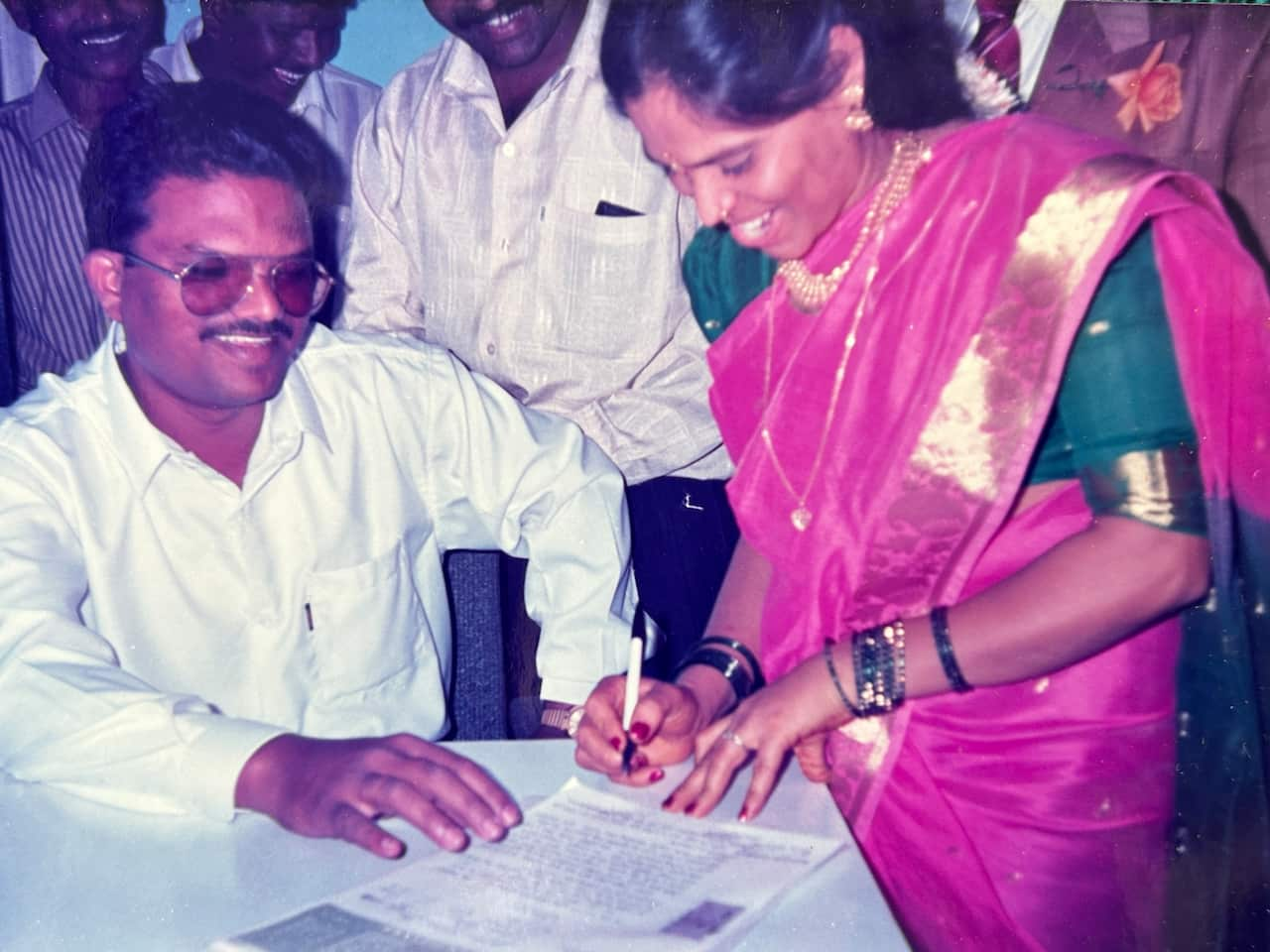 A man in a white shirt and thick glasses watches and smiles and a woman in a simple purple saree signs a marriage certificate with a white pen.