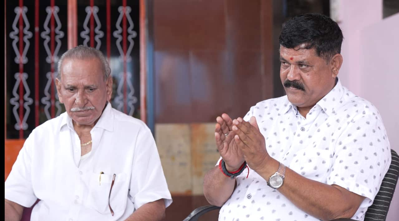 Two men wear white shirts and moustaches. On the right the man wearing a watch holds his hands together. 