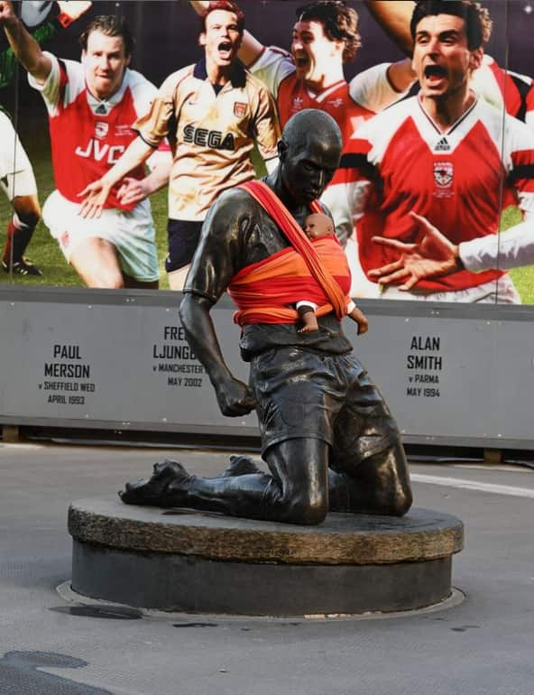 A statue of footballer Thierry Henry with a baby doll strapped to it in a red and orange sling.