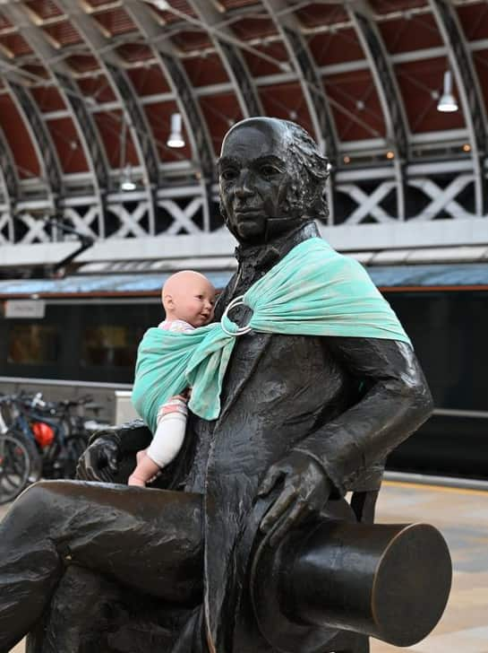 A statue of a man at a train station with a toy baby strapped to it using a blue sling.