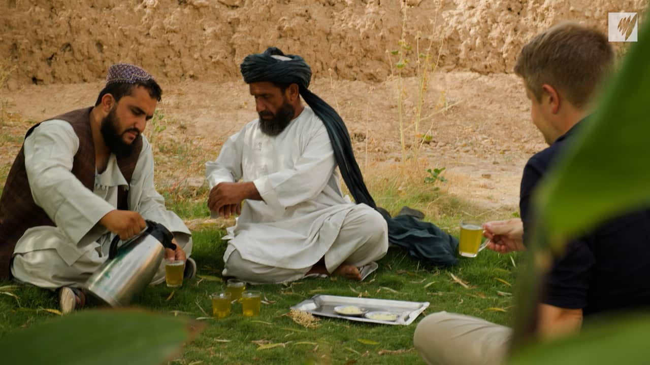 Gul Mohammad in green black turban and white robes sits next to a man pouring yellow tea