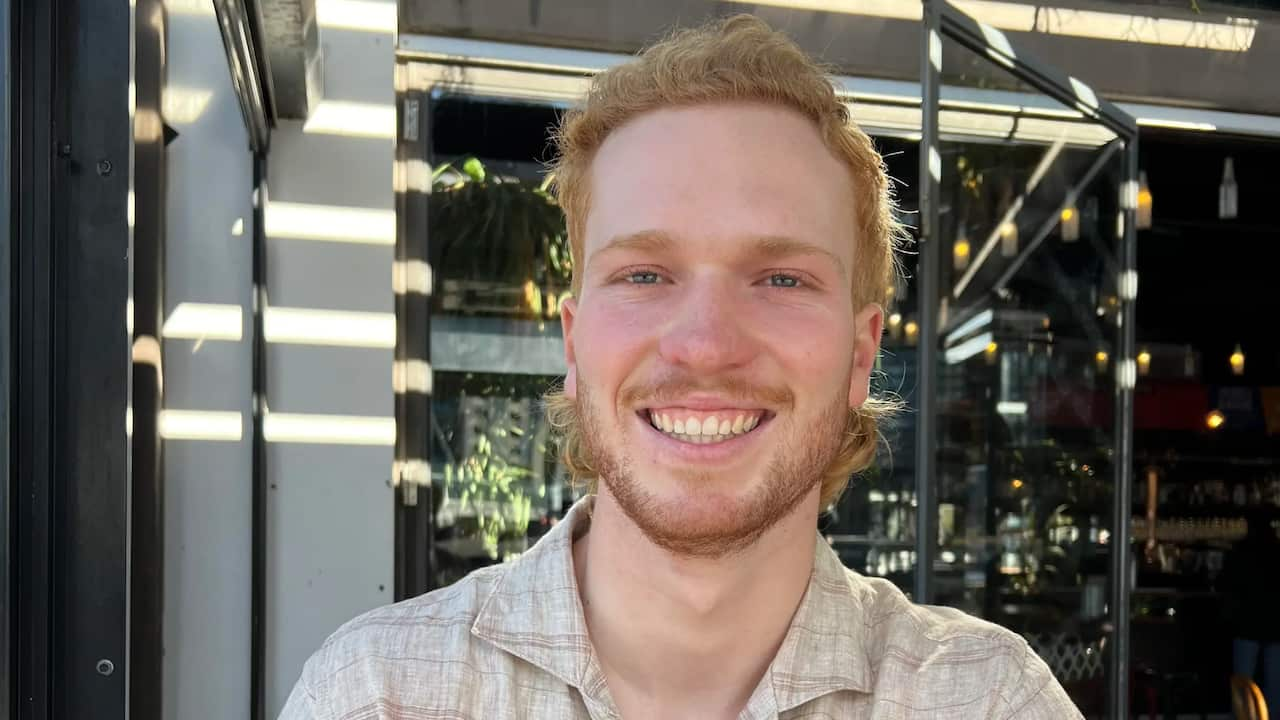 A man with ginger coloured long hair and a beard is wearing a summery shirt and holds a beer in a pub smiling.